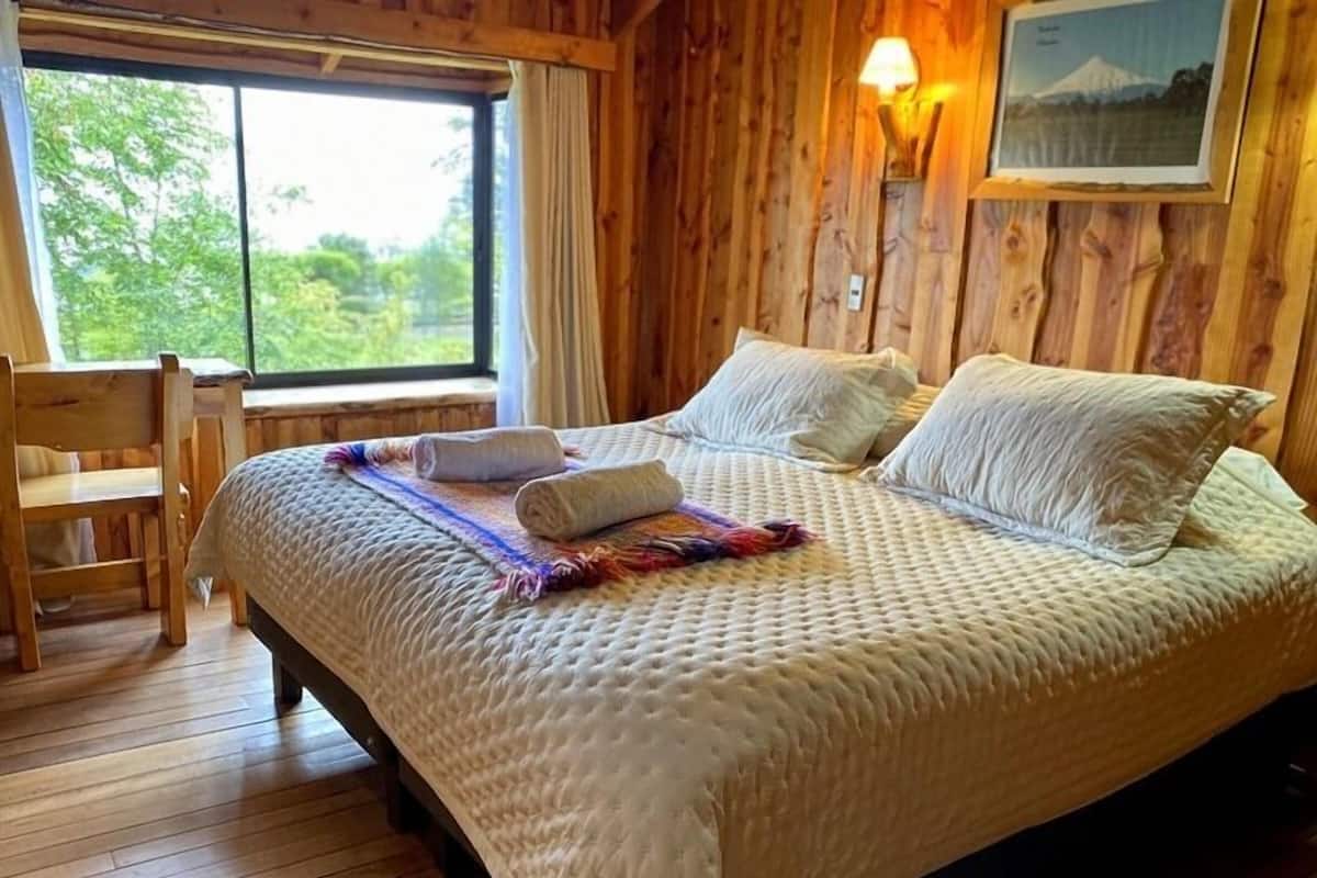 A wooden cabin bedroom with a bed, pillows, a chair, and a window with a view of trees.
