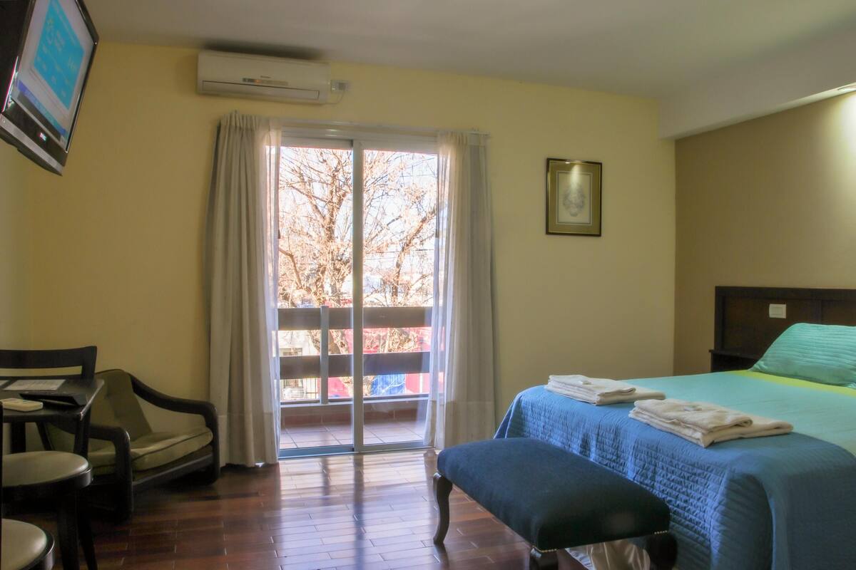 A hotel room with a bed, a desk, a chair, a window with curtains, and an air conditioning unit.