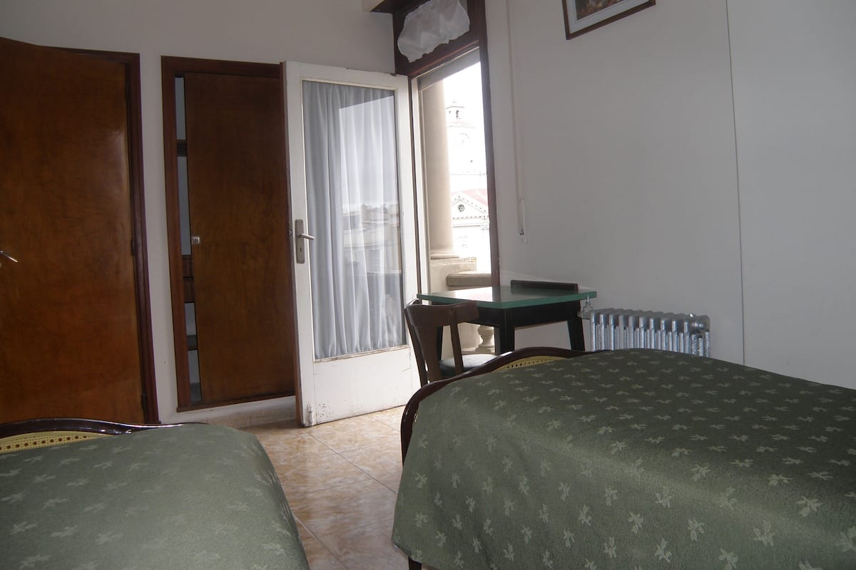 Une chambre avec deux lits, une table, une chaise, un radiateur et une porte donnant sur un balcon.