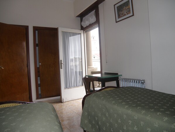 A room with two beds, a wooden door, a window with curtains, a table, and a radiator.