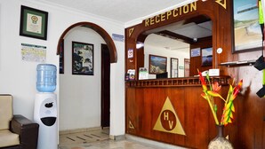 A hotel reception area with a wooden reception desk, a water dispenser, a waste bin, a reception mirror, and a vase with flowers. - Hotel Venecia Confort (San Juan de Pasto)