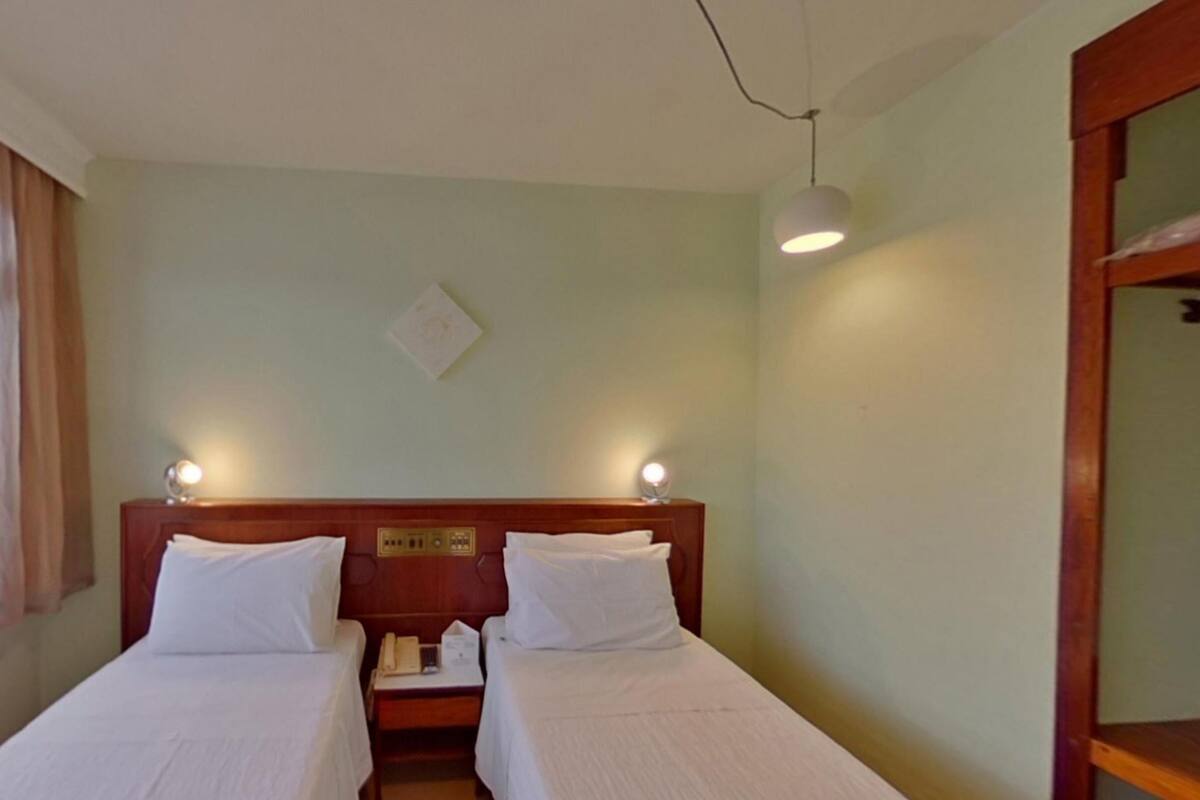 Ein Hotelzimmer mit zwei Einzelbetten, einem Nachttisch mit Lampe und einem Fenster mit durchsichtigen Vorhängen.