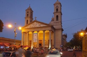 Un edificio histórico con dos torres y una entrada central, rodeado por una calle concurrida con gente y vehículos.