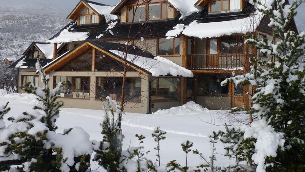 Ein großes, mehrstöckiges Chalet aus Holz mit schneebedeckten Dächern und Balkonen, umgeben von mit Schnee beladenen Bäumen.