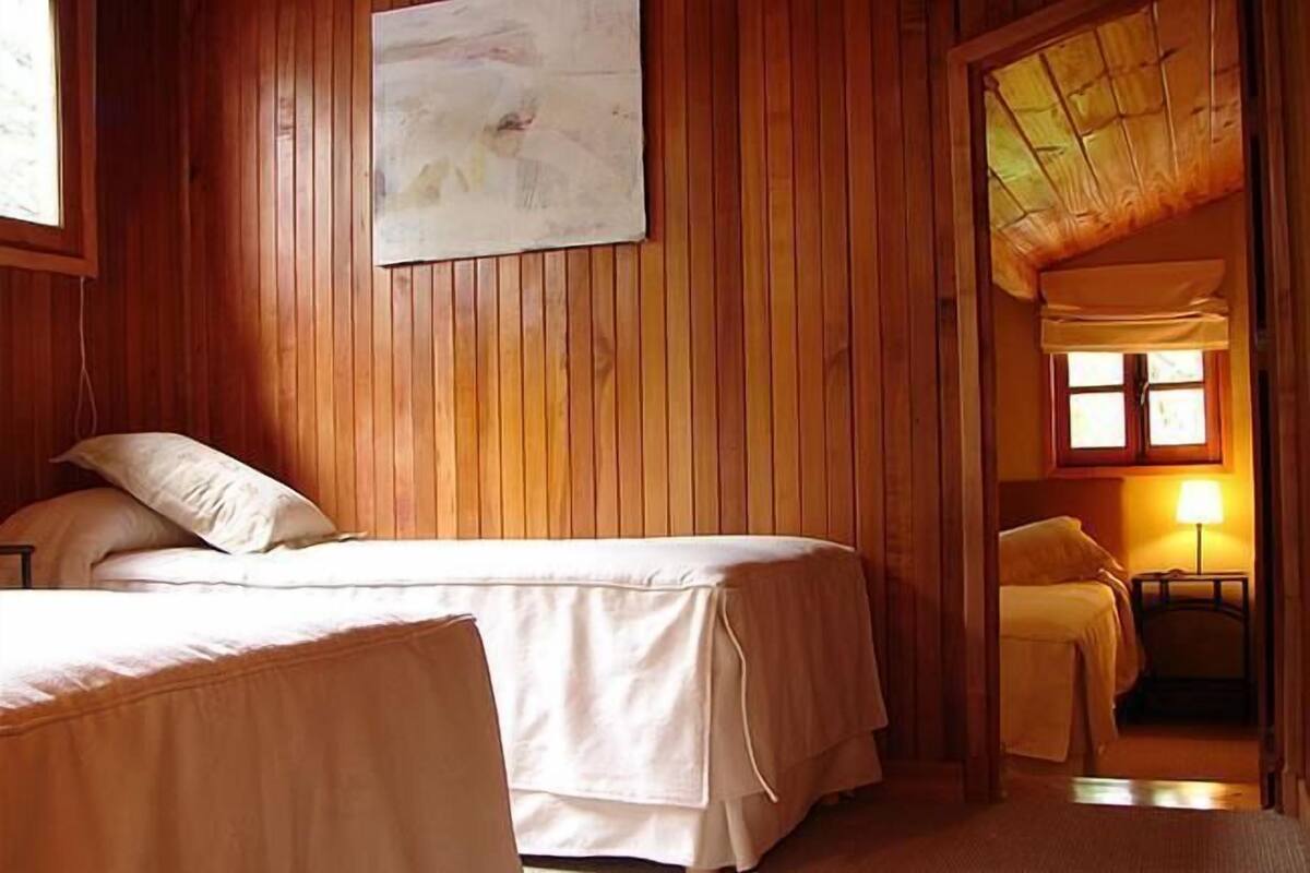 A room with wooden walls, two beds, a window with a view, and a lamp.