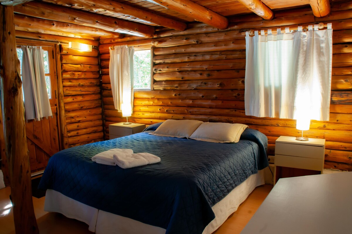 A log cabin bedroom with a bed, two bedside tables, and a window with curtains.