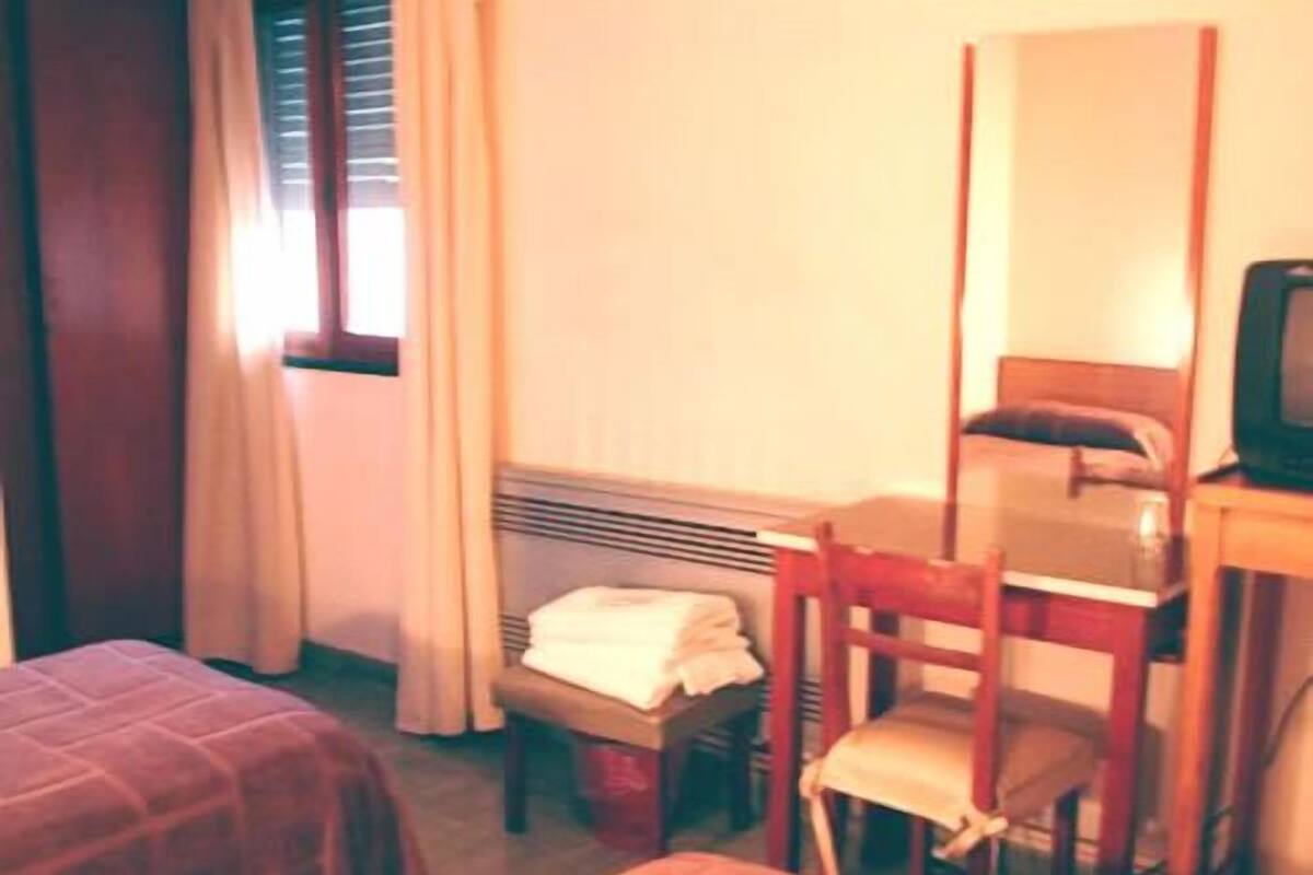 A small, compact room with a single bed, a chair, a desk with a television, and a window with curtains.
