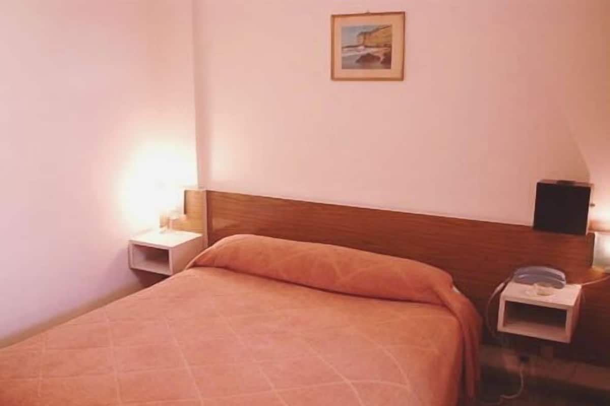 A single bed with a wooden headboard, a bedside table with a lamp, and a small shelf with a telephone.