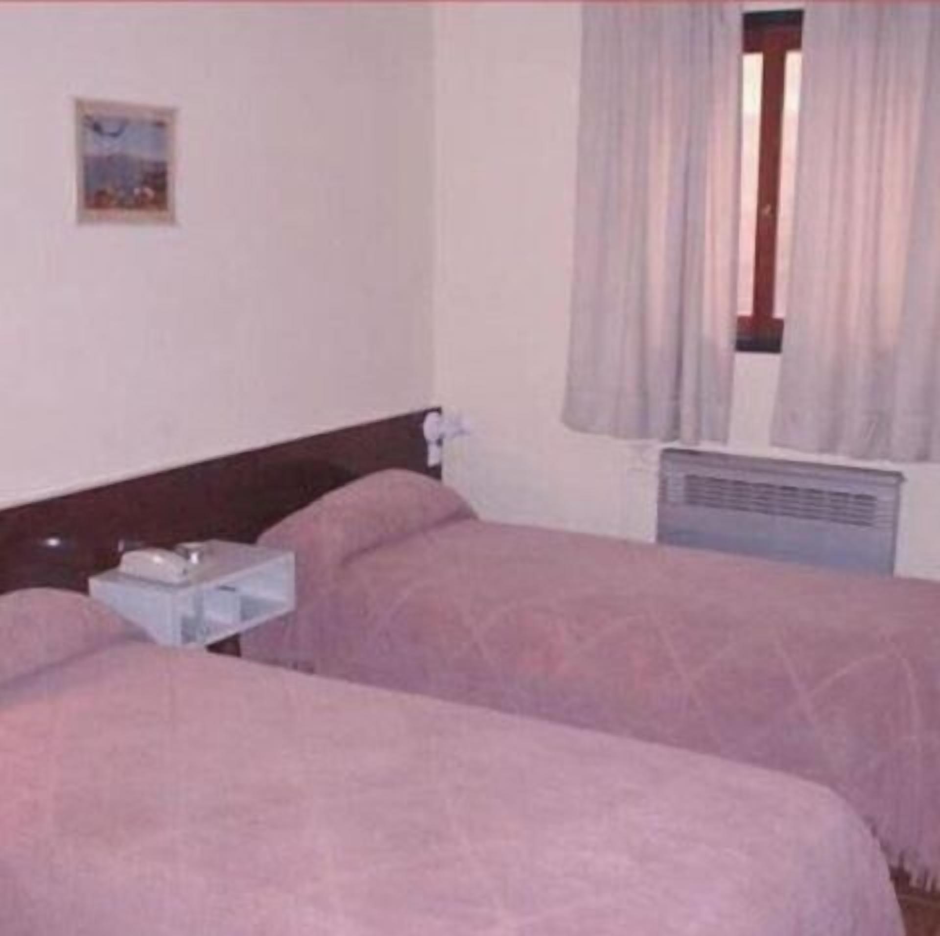 A hotel room with two beds, a small table, a nightstand, a window with curtains, and a wall-mounted air conditioning unit.