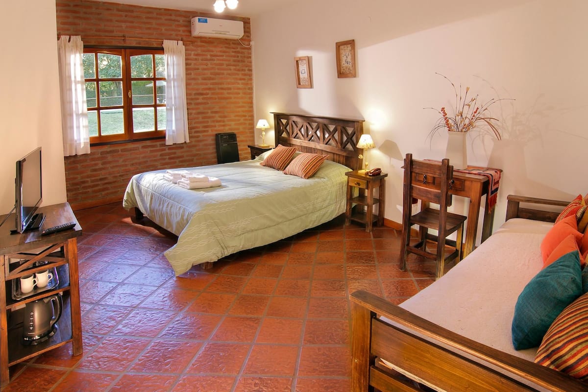 Een slaapkamer met een bed, een bureau, een stoel, een raam met gordijnen en een plant.