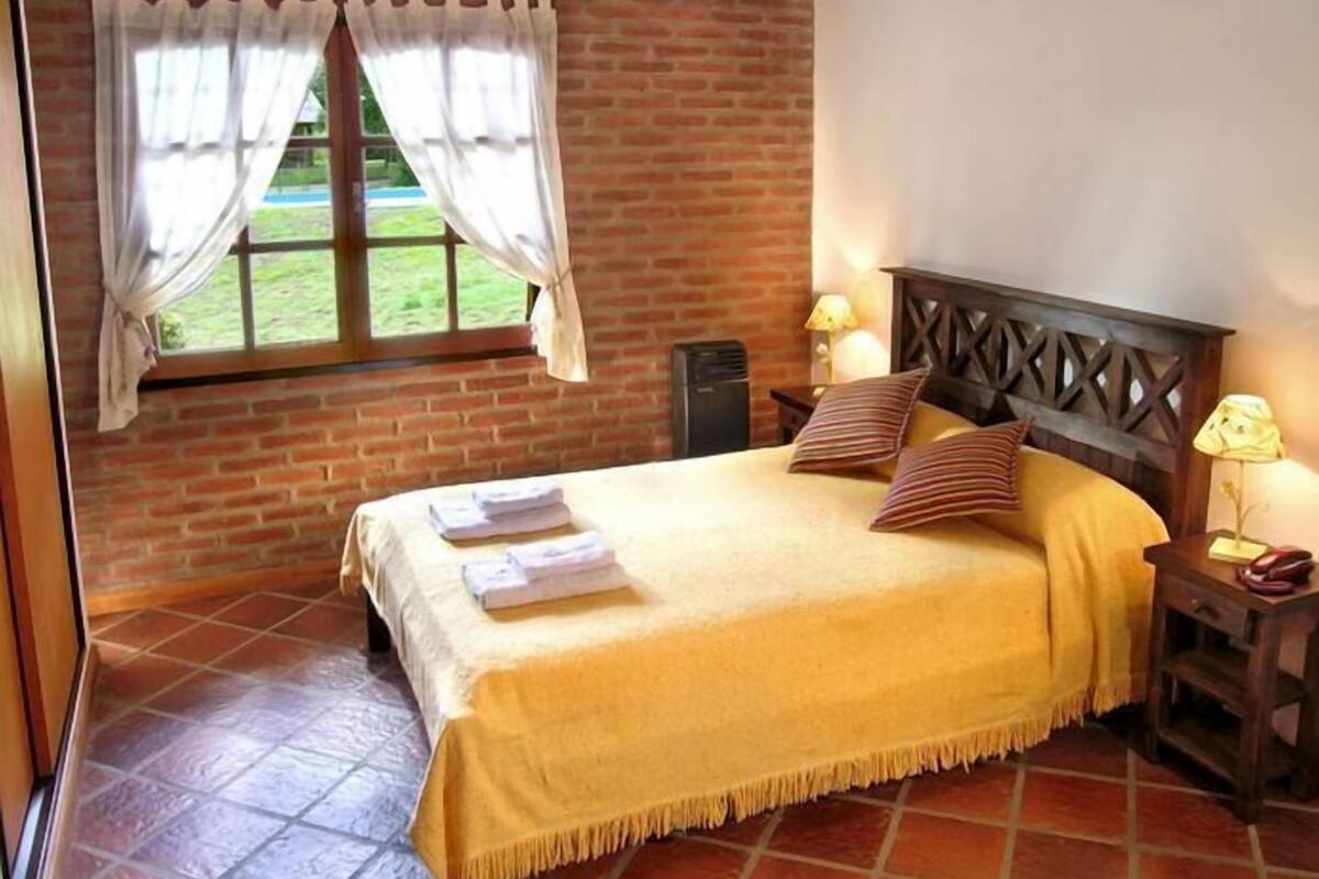 Een kamer met een bed, een raam met gordijnen, een lamp en een bakstenen muur.