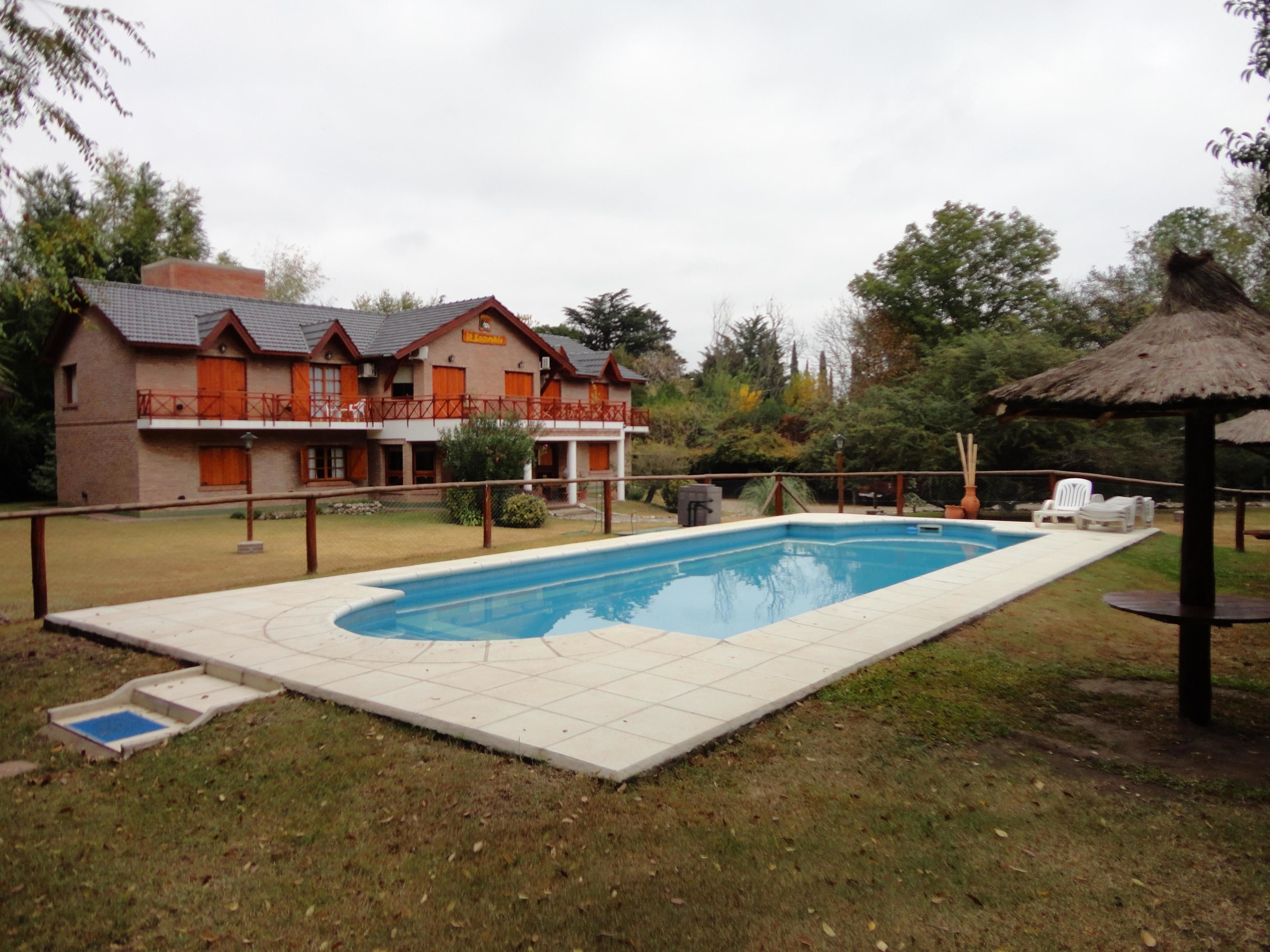Une piscine rectangulaire bordée d’une terrasse carrelée, entourée d’une pelouse et d’un bâtiment doté d’un toit rouge et de volets orange.
