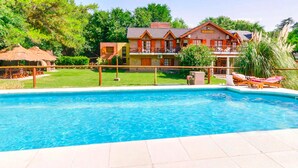 Una piscina con borde de baldosas, una casa con balcón y una construcción con techo de paja al fondo.