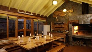 A rustic dining area with a long wooden table set for a meal, a stone fireplace, and a wooden ceiling with exposed beams. - Valldemossa Apart Hotel (San Martín de los Andes)