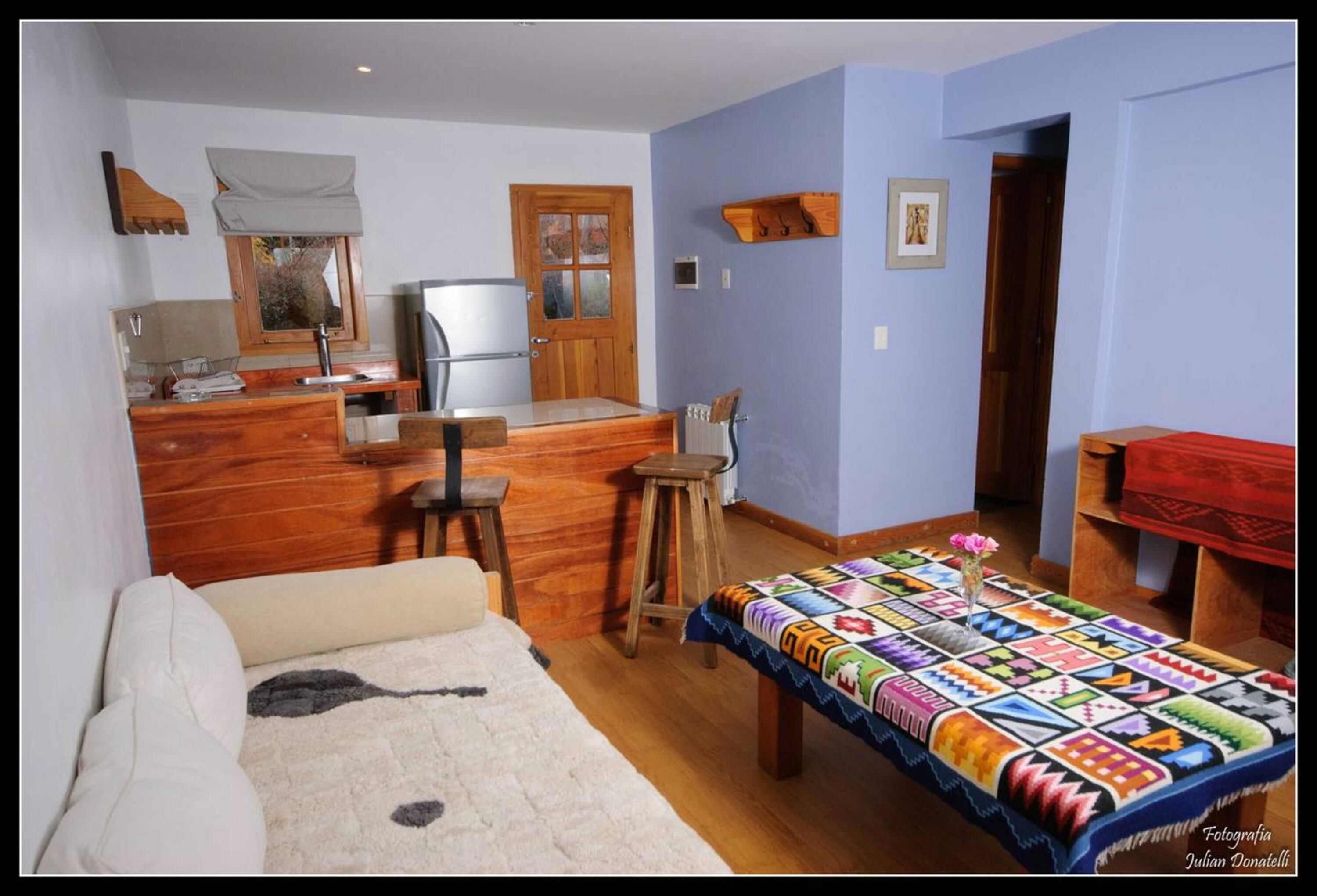Una habitación acogedora con un escritorio de madera, una silla, un taburete pequeño, una cama con una colcha blanca y gris estampada, y un colcha de colores en el suelo.