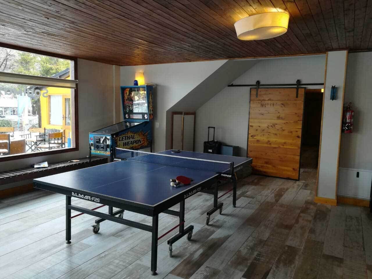 Una mesa de ping pong en una habitación con suelo de madera, una lámpara en el techo y vistas a través de una ventana.