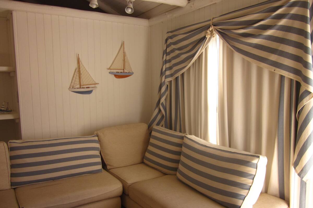 Um sofá de canto com almofadas listradas, uma mesinha com abajur e uma janela com cortinas listradas.