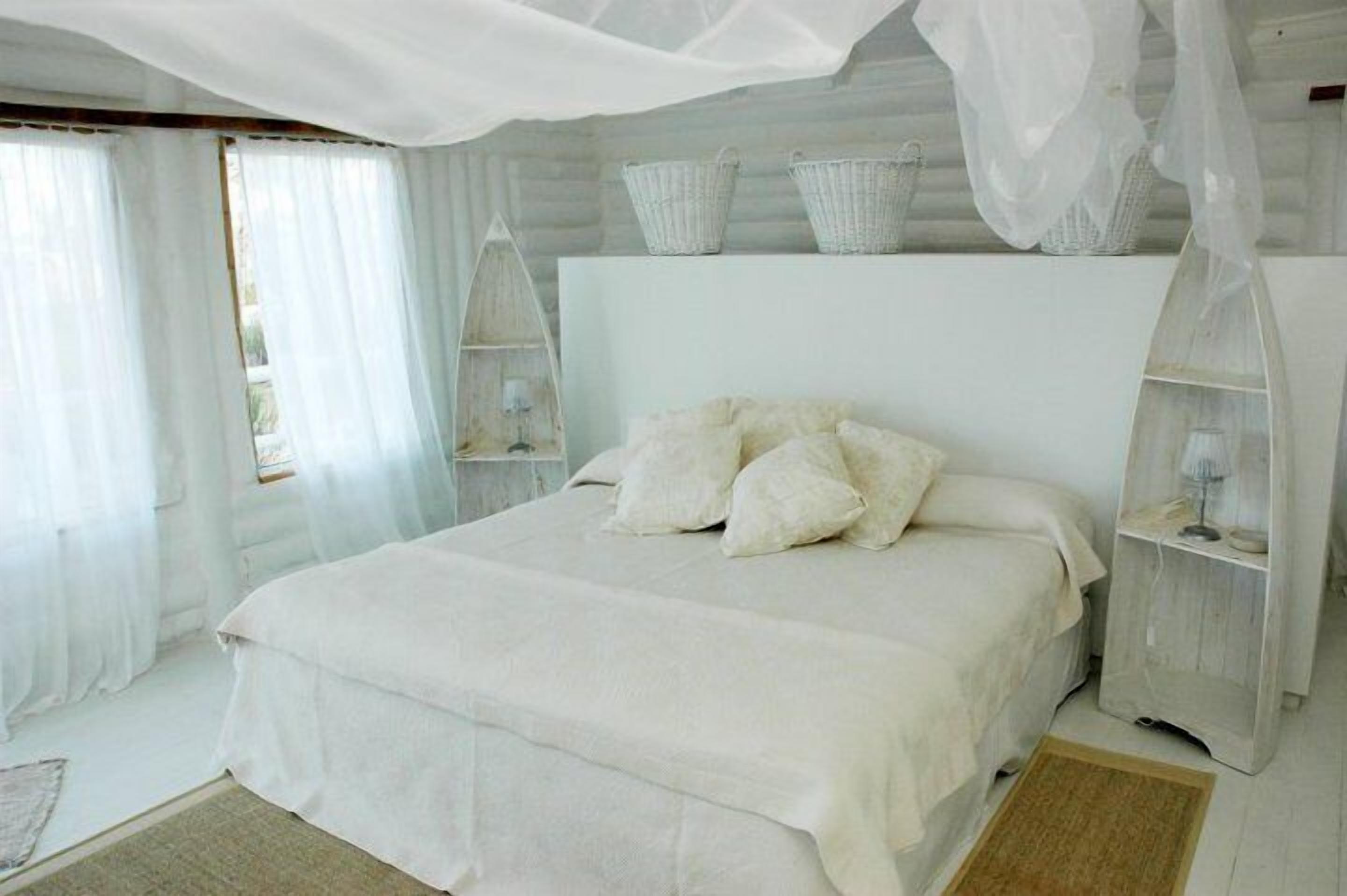 Un dormitorio con una cama grande, ropa de cama blanca, dosel y una mesita de noche de madera con cestas.