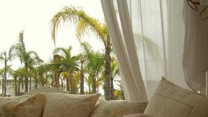 Une chambre avec un lit, une fenêtre ornée de rideaux légers et une vue sur les palmiers à l’extérieur.
