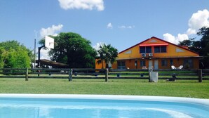 A swimming pool with a wooden deck in the foreground. In the background, there is a large orange building with a red roof, surrounded by trees and a clear blue sky. - Hotel Sanguinetti (Victoria)