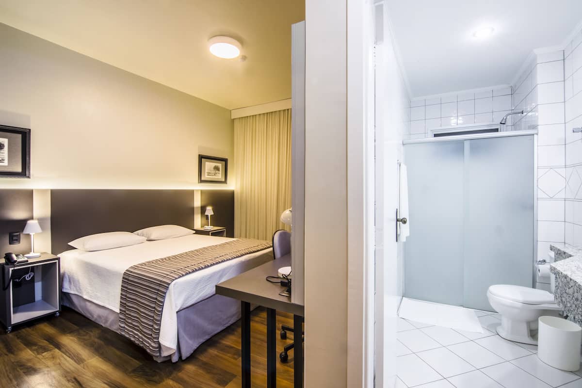 Habitación de hotel con cama, mesita de noche, lámpara y vistas al baño.