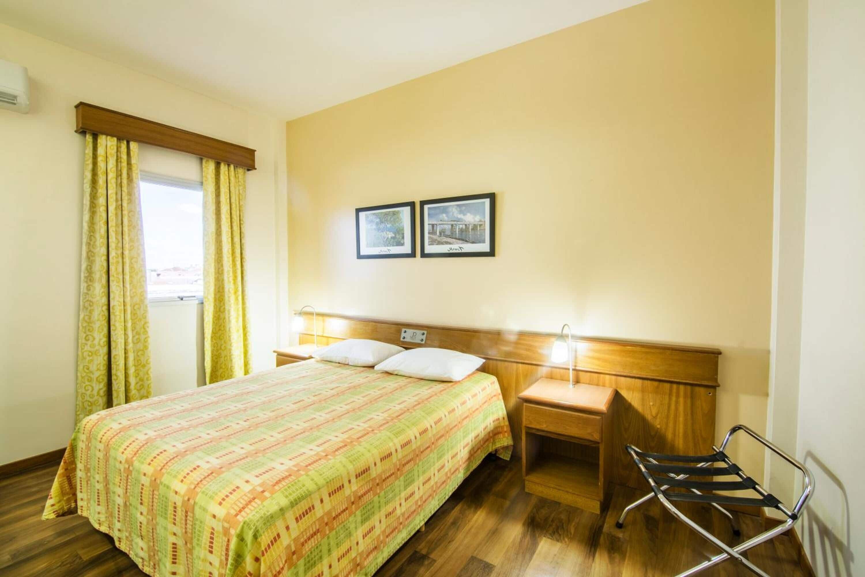A hotel room with a bed, wooden headboard, two bedside tables, a chair, and two framed pictures on the wall.