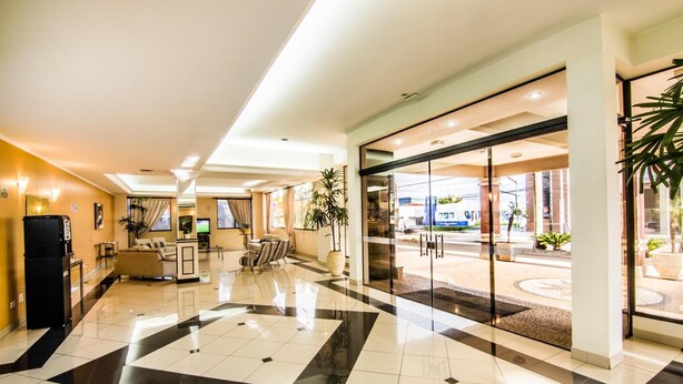 A hotel lobby with a check-in counter, seating area, and a glass door leading outside. - JR Hotel (Marilia)