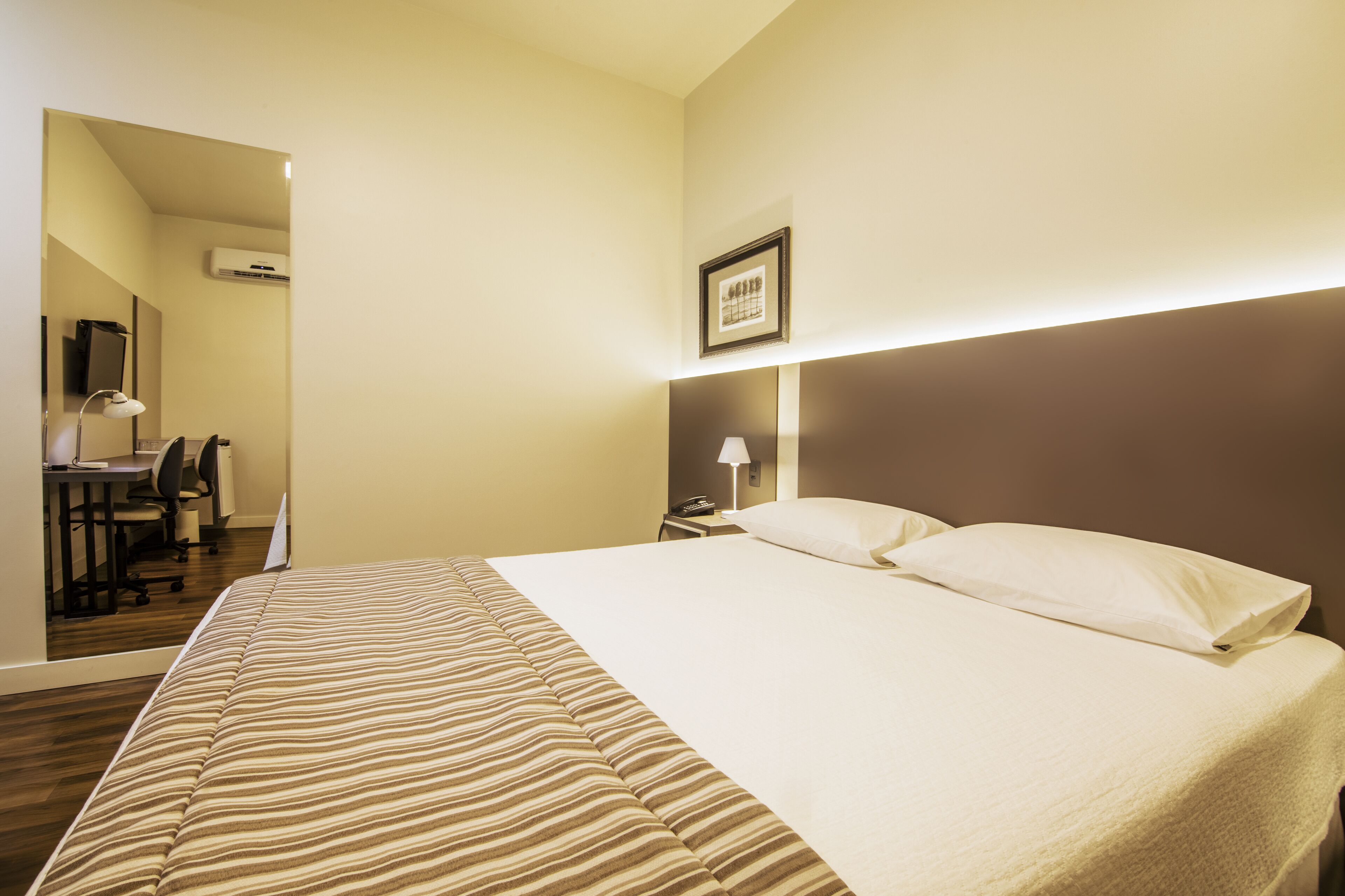 A hotel room with a bed, a desk, and a chair. There is a framed picture on the wall and a lamp on the nightstand.