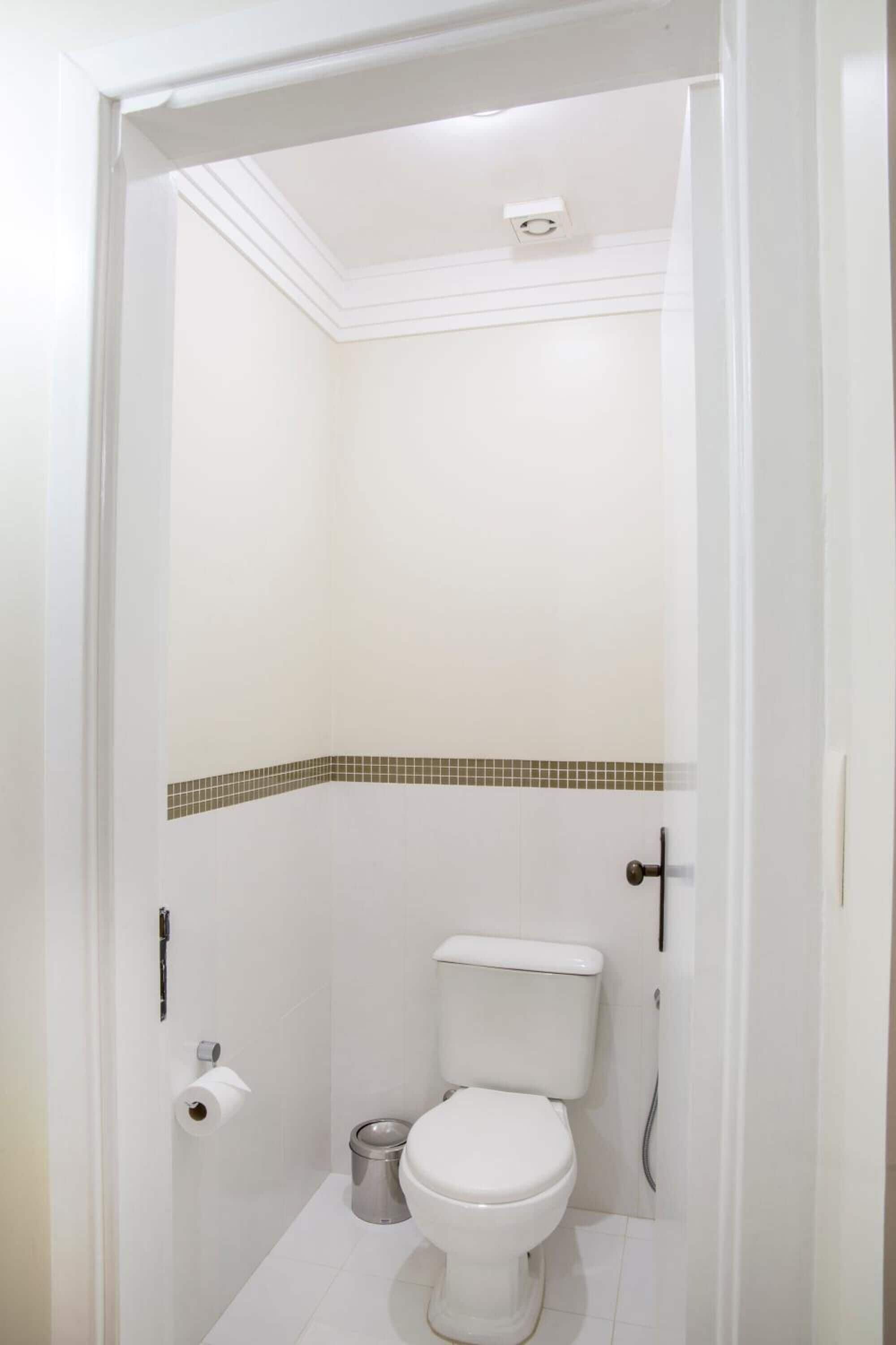 Une petite salle de bain blanche, avec des toilettes, une poubelle et un rouleau de papier toilette accroché au mur.