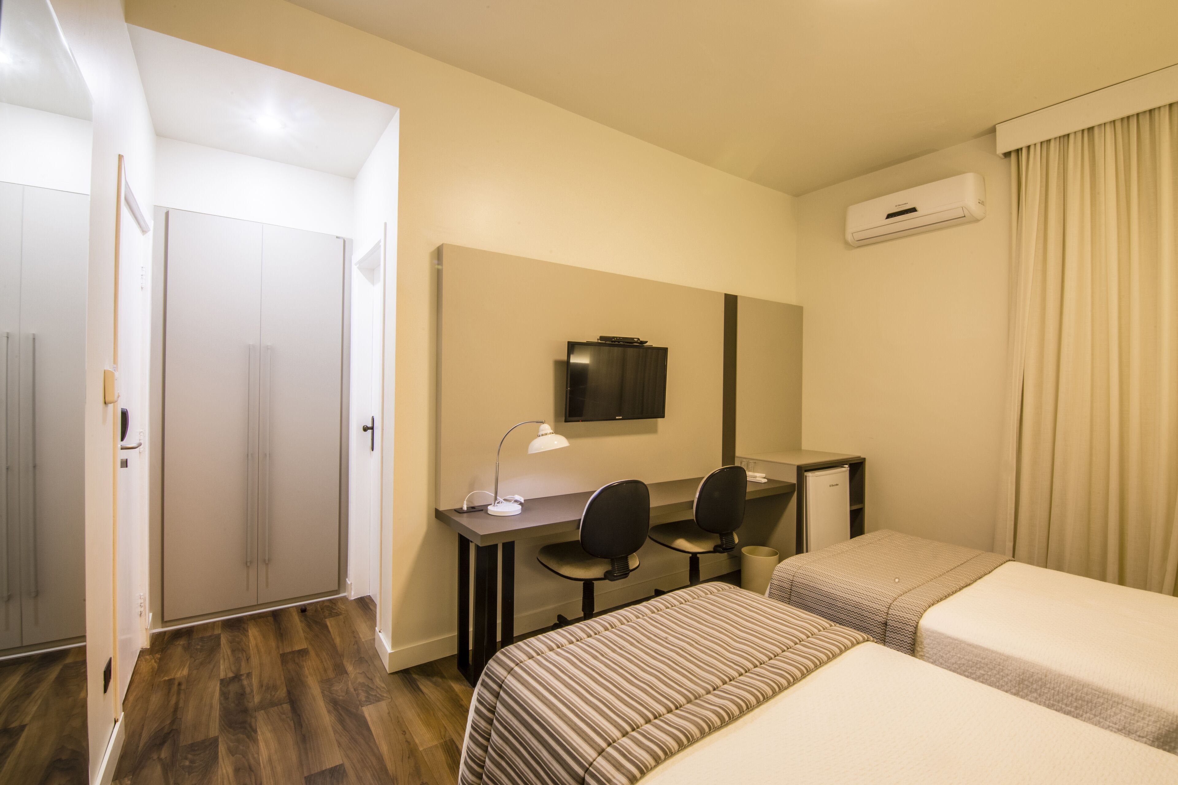 Una habitación de hotel con dos camas individuales, un escritorio con una silla, un televisor y un aire acondicionado.