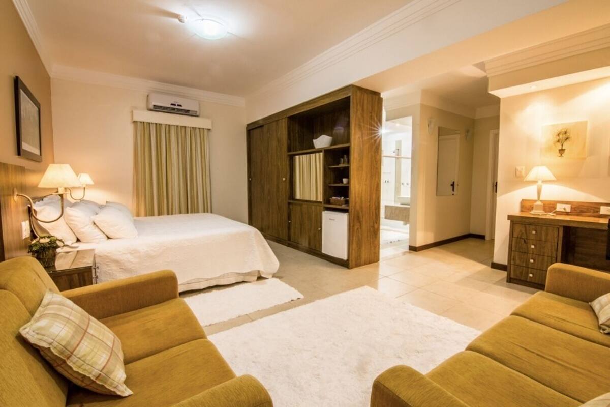 Habitación de hotel con cama, dos sofás, un armario de madera, un escritorio con lámpara y baño a la vista a través de una puerta abierta.