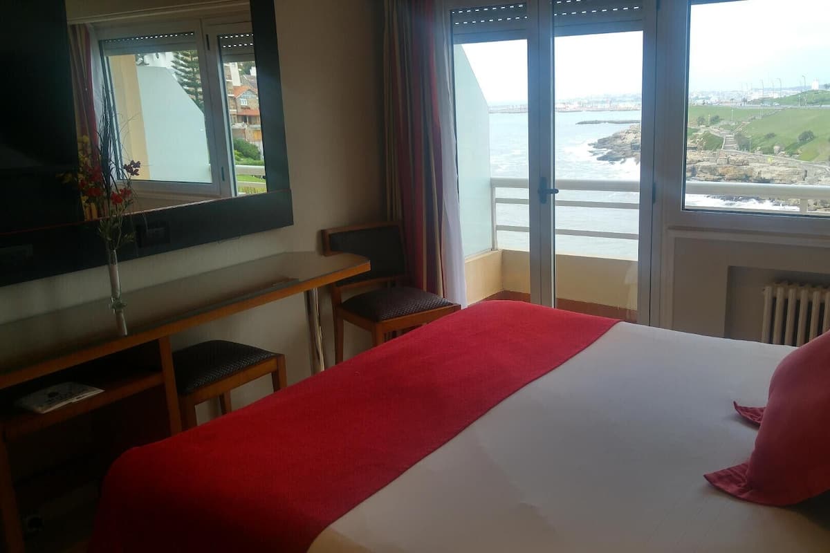 A hotel room with a bed, a desk, a chair, a mirror, and a view of the sea and cityscape.