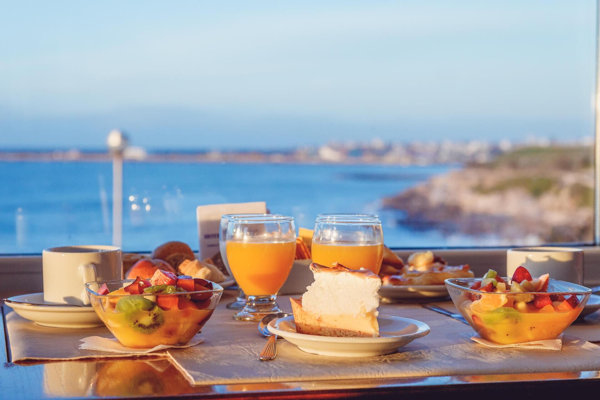 A table with a fruit bowl, a glass of orange juice, and a slice of cheesecake, with a view of the sea in the background.