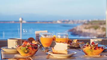 A table with a fruit bowl, a glass of orange juice, and a slice of cheesecake, with a view of the sea in the background.