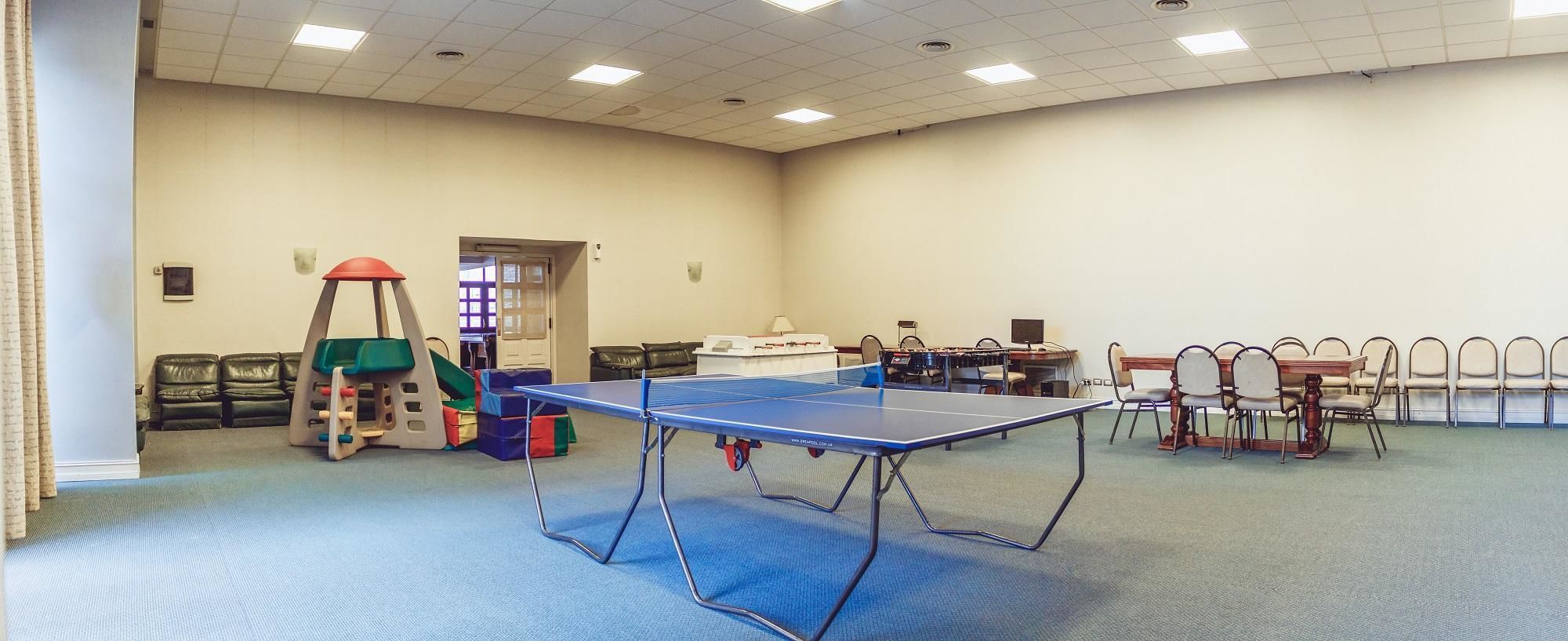 Una habitación amplia con mesa de ping pong, sillas y una pequeña zona de juegos con un tobogán.