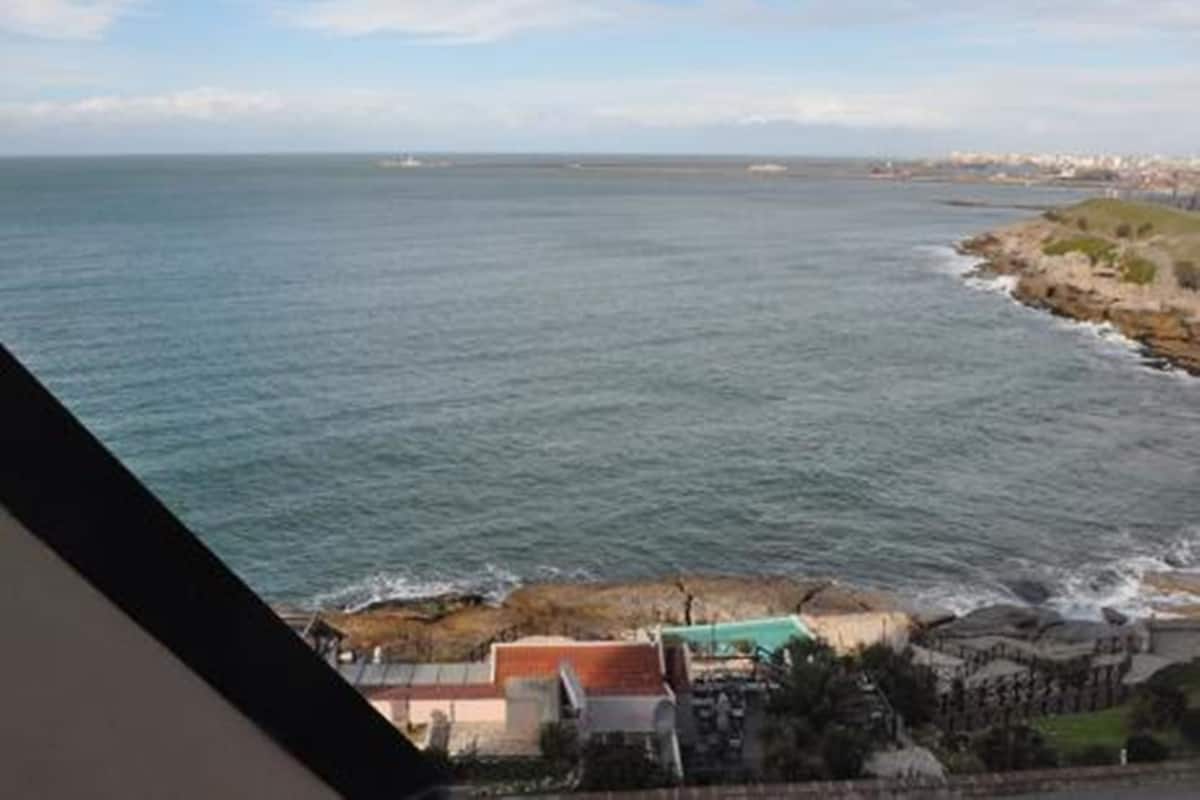 A coastal view with a rocky shoreline, a building with a red roof, and a clear sky.