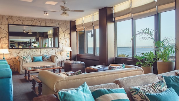 A spacious living room with a large window offering a view of the sea, a stone wall, and a ceiling fan. - Hotel Las Rocas (Mar del Plata)