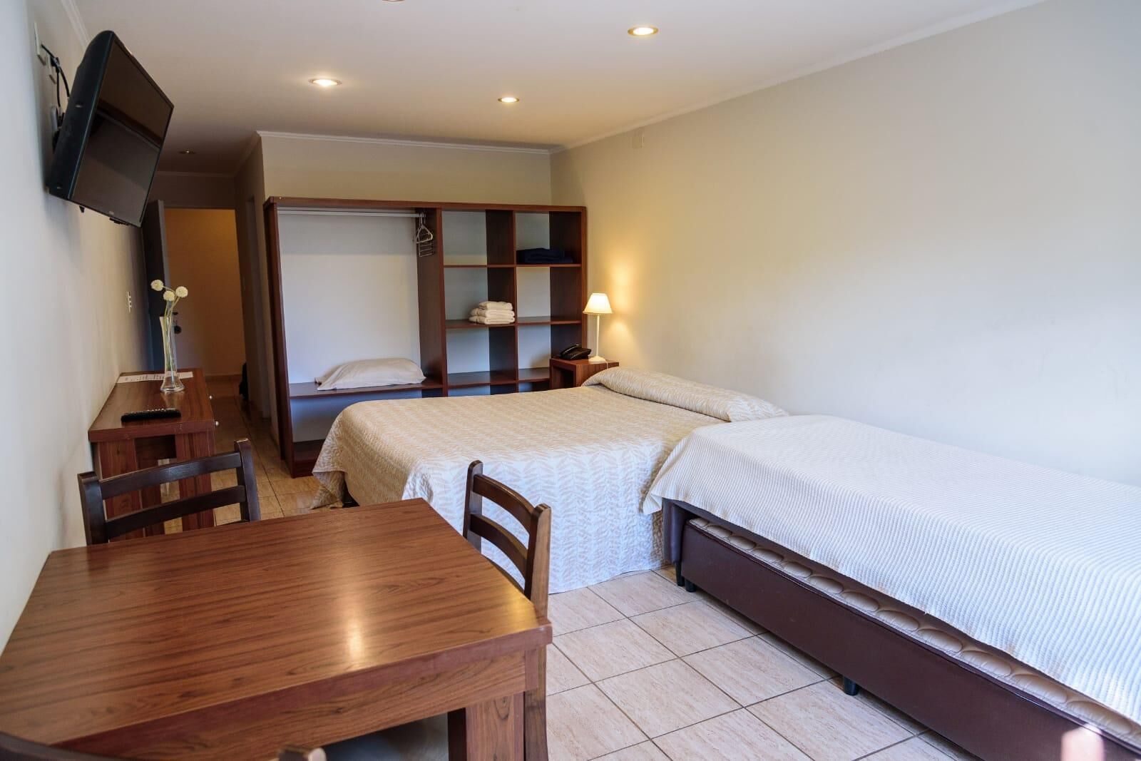 A small, well-lit room with two single beds, a wooden table, a TV mounted on the wall, and a wardrobe with shelves.