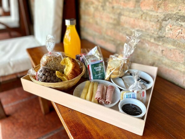 Bandeja de desayuno con pan, bollos, una botella de zumo de naranja y salsas sobre una mesa de madera frente a un muro de ladrillo.
