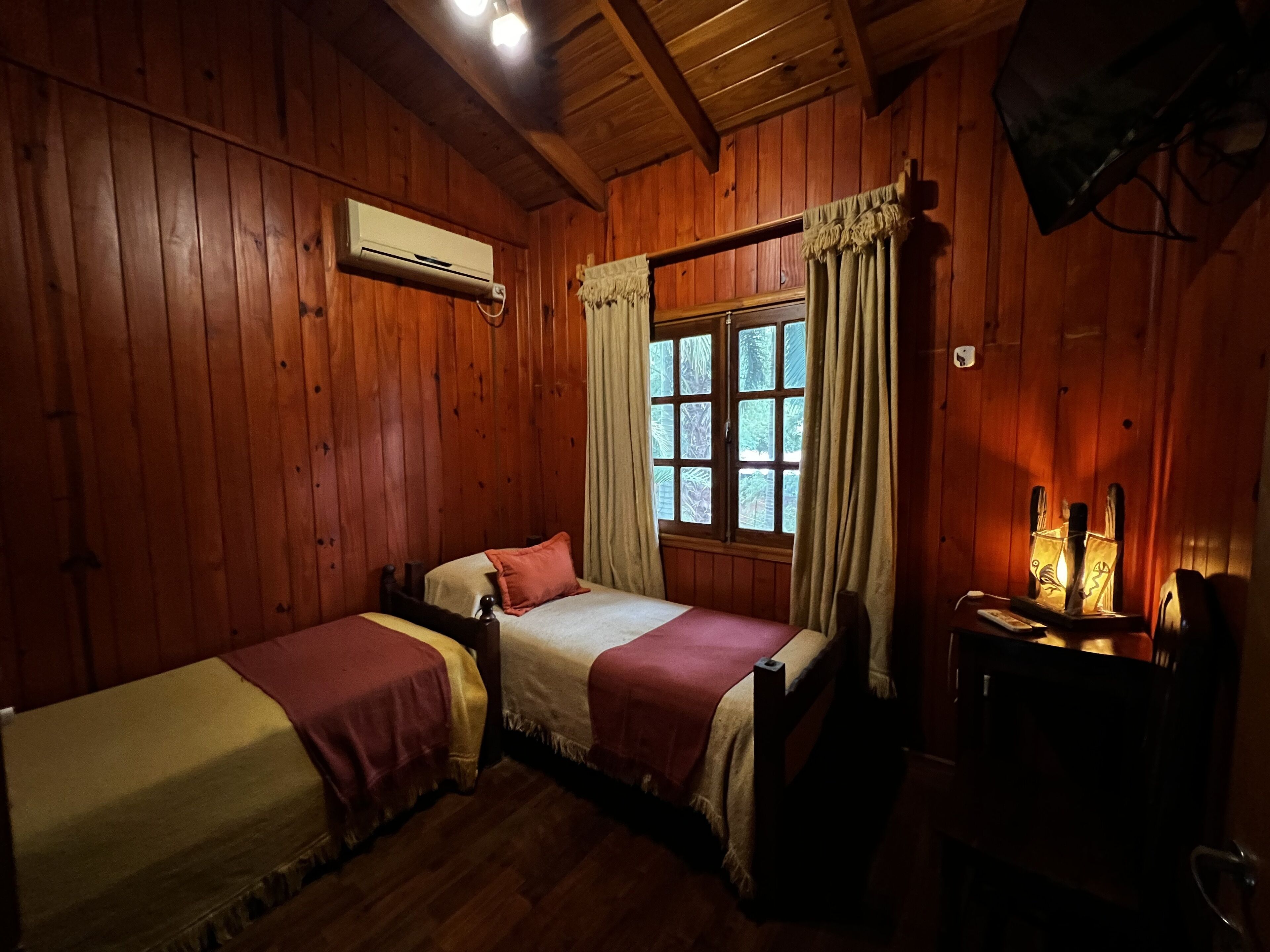 A wooden cabin room with two beds, a window with curtains, a wall-mounted air conditioner, and a table with candles.