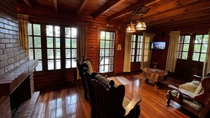 Interior de una cabaña de troncos con chimenea, muebles de madera, ventanas amplias y un televisor.