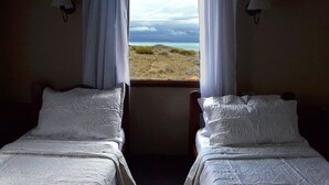 Ein Schlafzimmer mit zwei Betten, weißer Bettwäsche und Blick auf einen Strand durch ein Fenster.