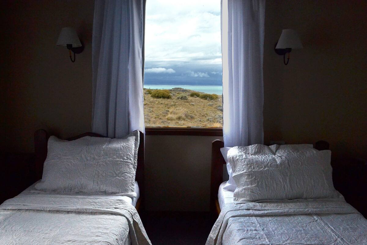 Quarto com duas camas, roupa de cama branca e vista para a praia pela janela.