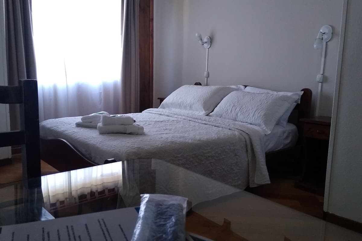 Uma cama bem arrumada com lençóis brancos, uma mesinha de cabeceira com abajur e uma janela com cortinas.