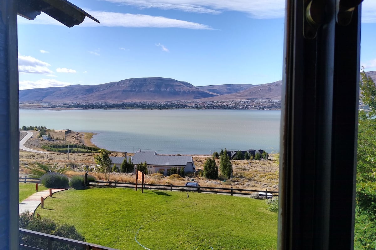 Vista de um lago com montanhas ao fundo, uma área gramada em primeiro plano e um edifício com telhado escuro.