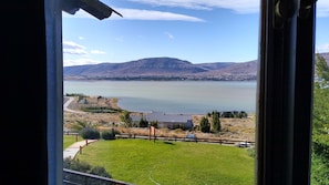 A view of a lake with mountains in the background, a grassy area in the foreground, and a building with a dark roof.