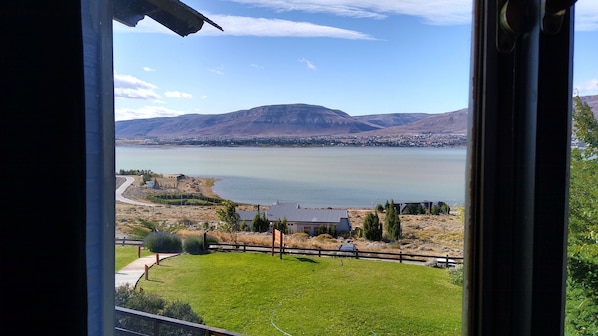 A view of a lake with mountains in the background, a grassy area in the foreground, and a building with a dark roof.
