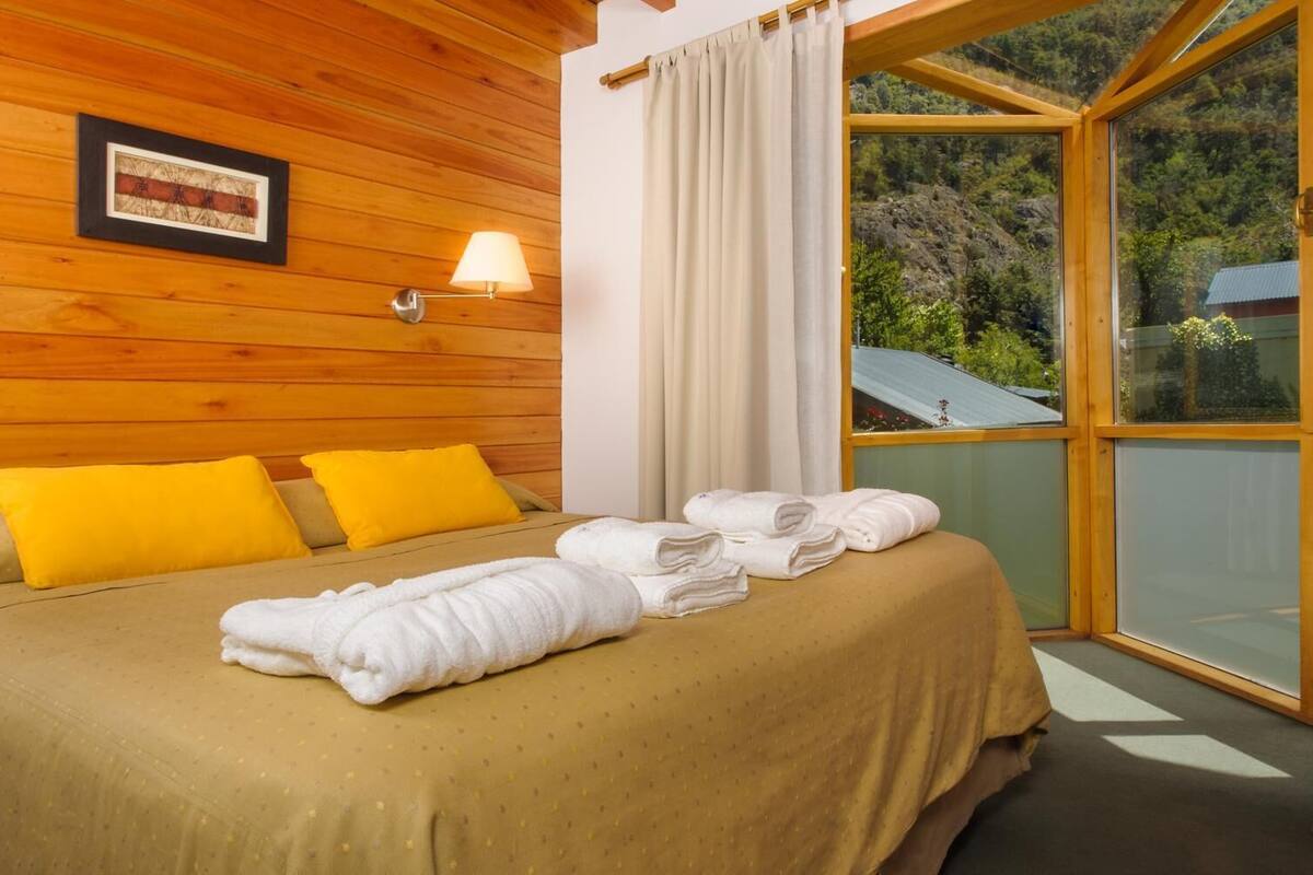 A wooden room with a bed, yellow pillows, a window with curtains, and a towel on the bed.