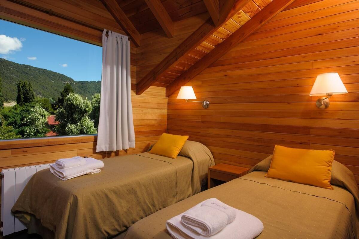 A wooden cabin room with two beds, each with a pillow and a rolled towel. There are two wall-mounted lamps and a window with a curtain.
