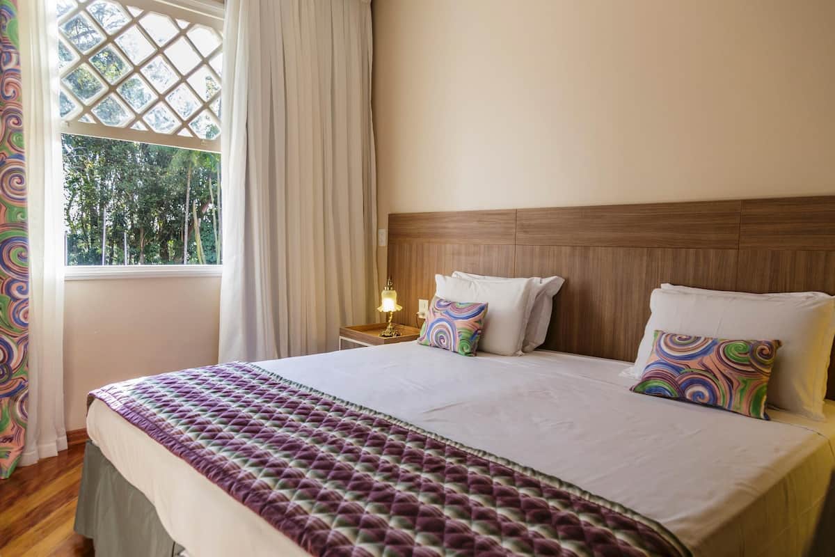 Una cama bien tendida con una colcha estampada, dos almohadas, un cabecero de madera, una mesita de noche con una lámpara y una ventana con cortinas.